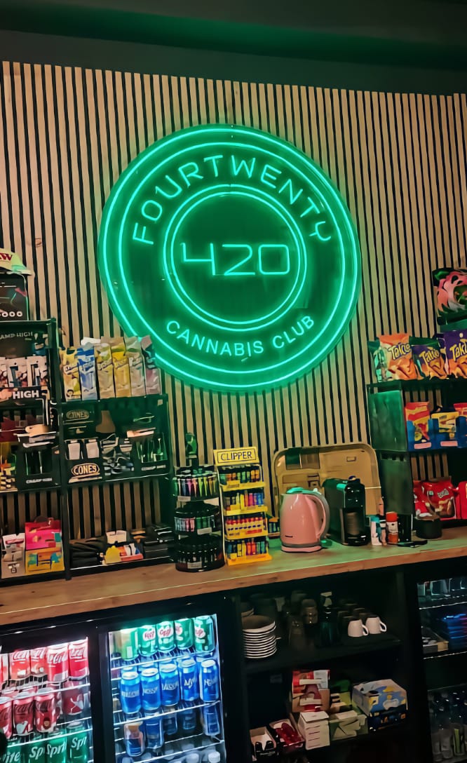 Un letrero de ne&oacute;n verde brillante con el logotipo del club de cannabis Four Twenty en Valencia, situado sobre una barra de bar llena de aperitivos, bebidas y encendedores Clipper.