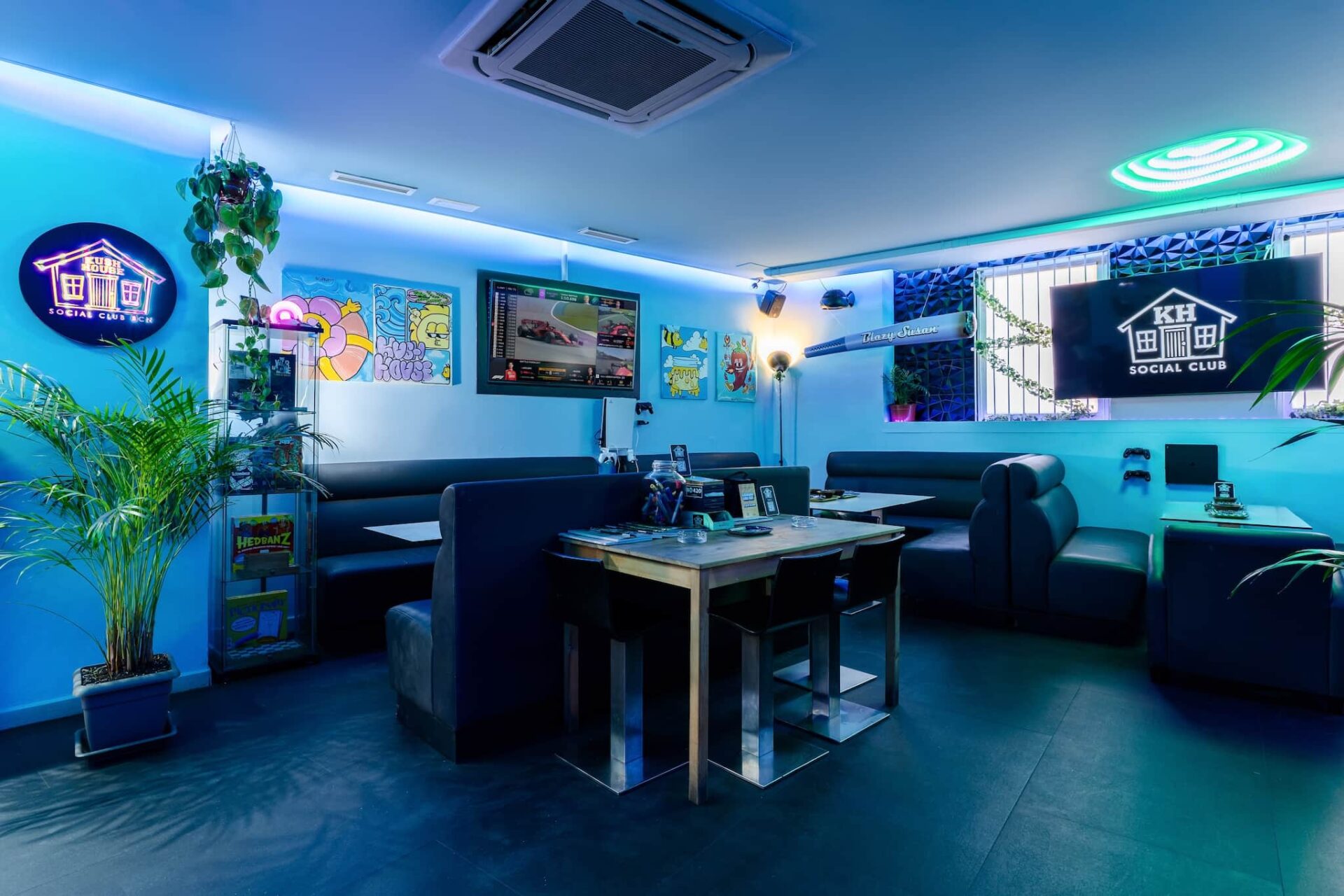 Interior del Kush House Social Club de Barcelona, con iluminaci&oacute;n ambiental azul, asientos tipo sof&aacute;, pantallas para juegos, coloridas obras en la pared y plantas en maceta
