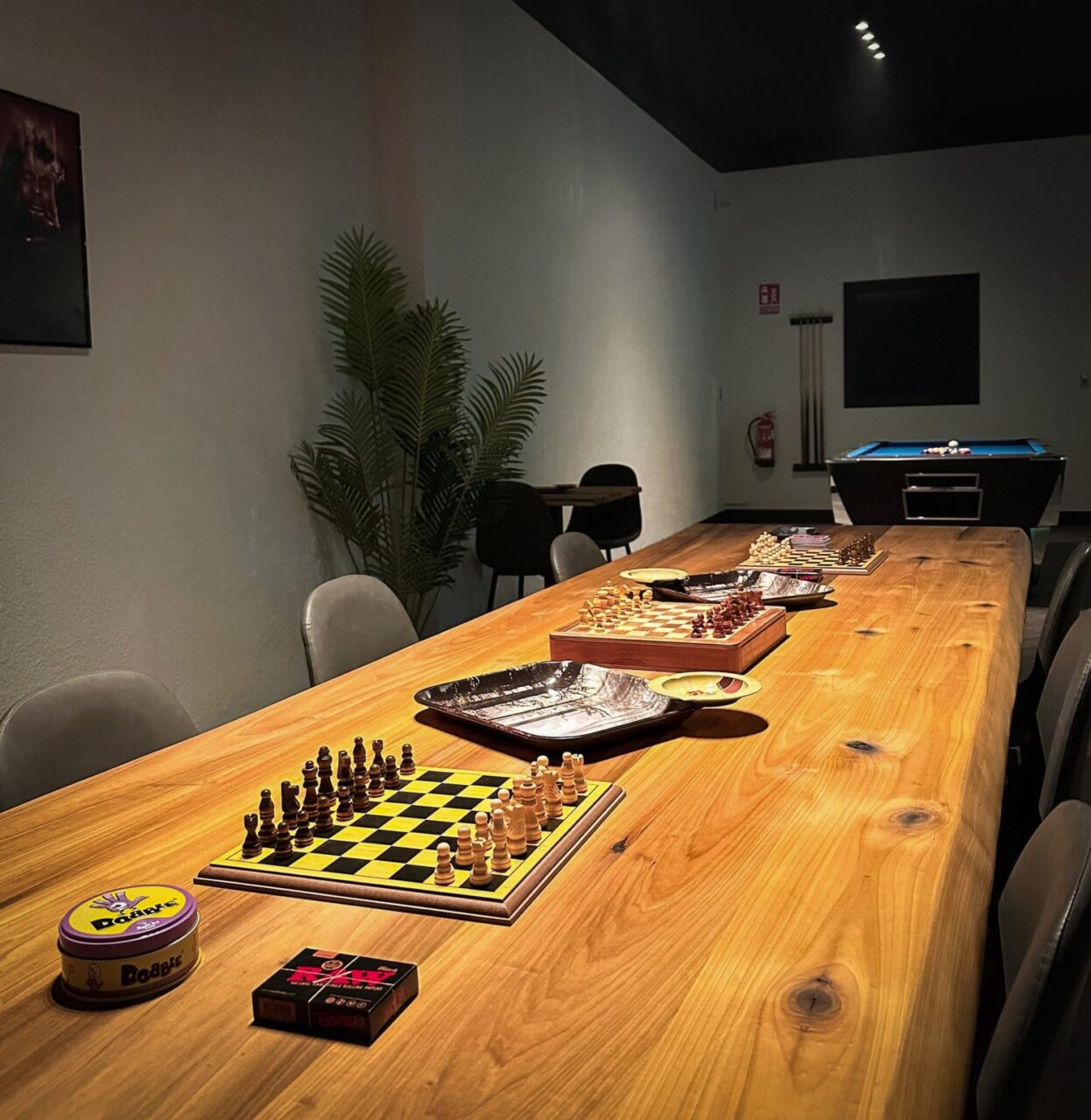 Una larga mesa de madera con tableros de ajedrez, juegos de mesa y accesorios para fumar en el club de cannabis Four Twenty en Valencia, con una mesa de billar visible al fondo.