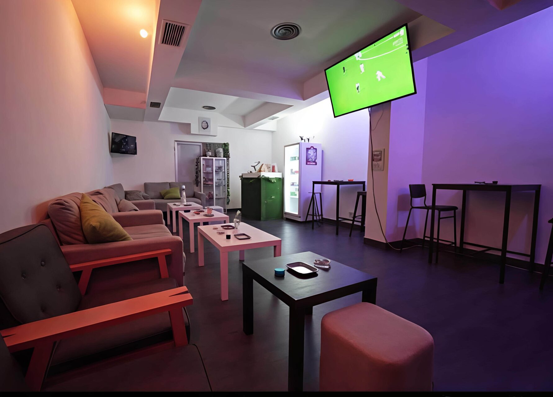 Zona de sal&oacute;n en el cannabis club Valhalla en Madrid con asientos, mesas altas, un refrigerador y un televisor transmitiendo un partido de f&uacute;tbol.